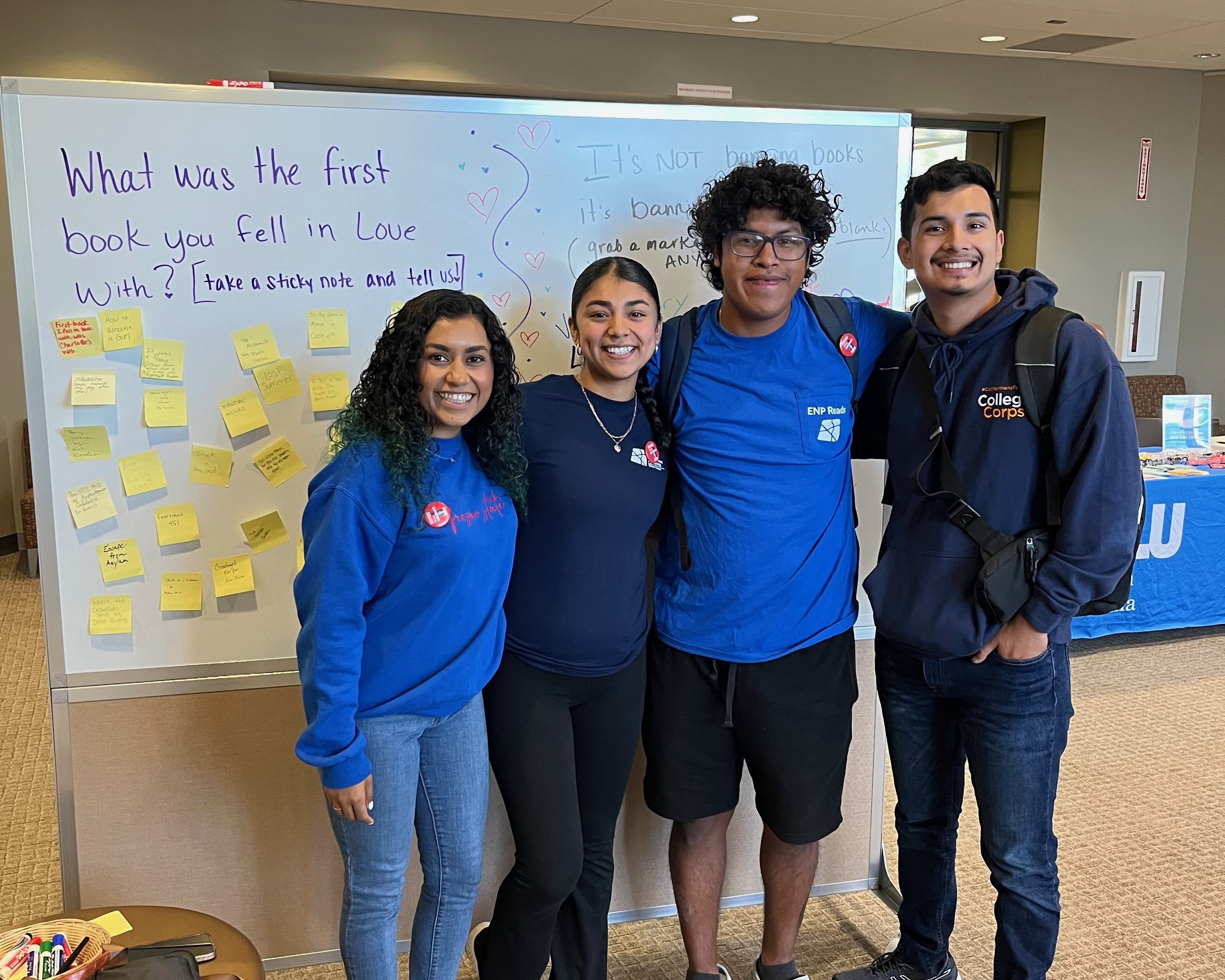Four students standing together in front of a whiteboard with post-it notes on it. The whiteboard has the question written on it: "What was the first book you fell in love with?"